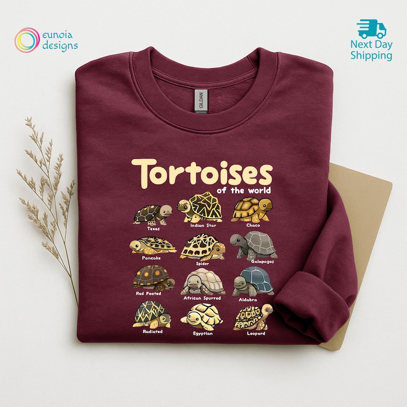 May include: Burgundy sweatshirt featuring the text "Tortoises of the World" above illustrations of various tortoise species. The sweatshirt has a crew neck and long sleeves. The design includes the names of each tortoise species.