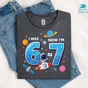 Puede incluir: Camiseta gris oscuro con un gr&aacute;fico colorido con tem&aacute;tica espacial. El dise&ntilde;o incluye el texto "I WAS 6 NOW I'M 7" con los n&uacute;meros azules 6 y 7, un astronauta en un cohete, planetas y estrellas. La camiseta est&aacute; sobre unos vaqueros azules.