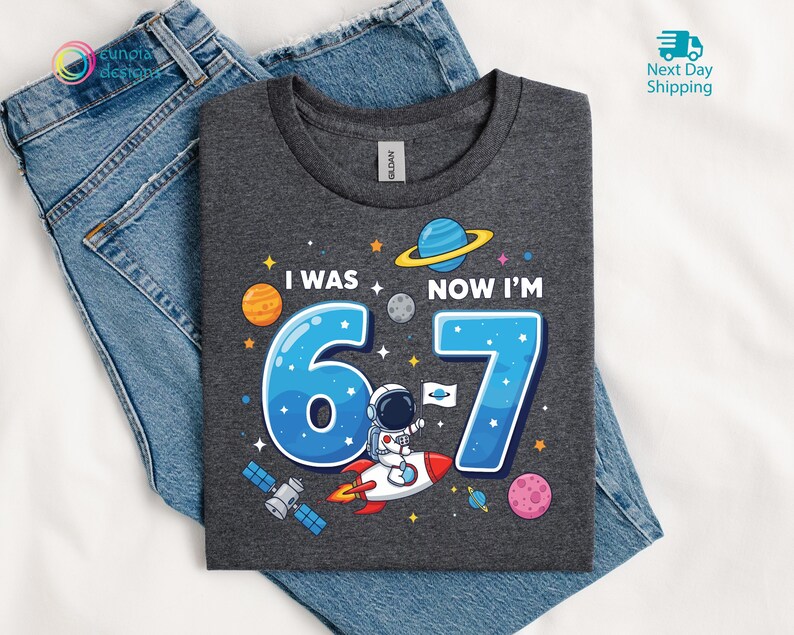 Puede incluir: Camiseta gris oscuro con un gr&aacute;fico colorido con tem&aacute;tica espacial. El dise&ntilde;o incluye el texto "I WAS 6 NOW I'M 7" con los n&uacute;meros azules 6 y 7, un astronauta en un cohete, planetas y estrellas. La camiseta est&aacute; sobre unos vaqueros azules.