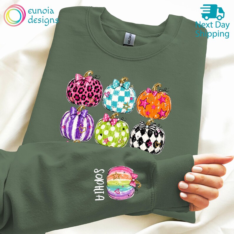Puede incluir: Una sudadera de cuello redondo color verde oliva con un gr&aacute;fico de seis calabazas estilizadas con varios patrones, incluyendo estampado de leopardo, cuadros y lunares, cada una con un lazo. La manga muestra una calabaza arco&iacute;ris y el nombre Sophia.