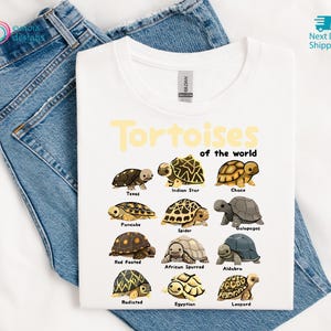 May include: White t-shirt featuring a colorful graphic of various tortoise species with their names. The text "Tortoises of the world" is printed in yellow above the illustrations. The shirt is displayed on a pair of blue jeans.