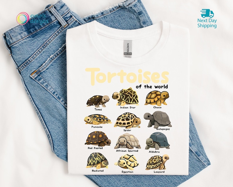 May include: White t-shirt featuring a colorful graphic of various tortoise species with their names. The text "Tortoises of the world" is printed in yellow above the illustrations. The shirt is displayed on a pair of blue jeans.