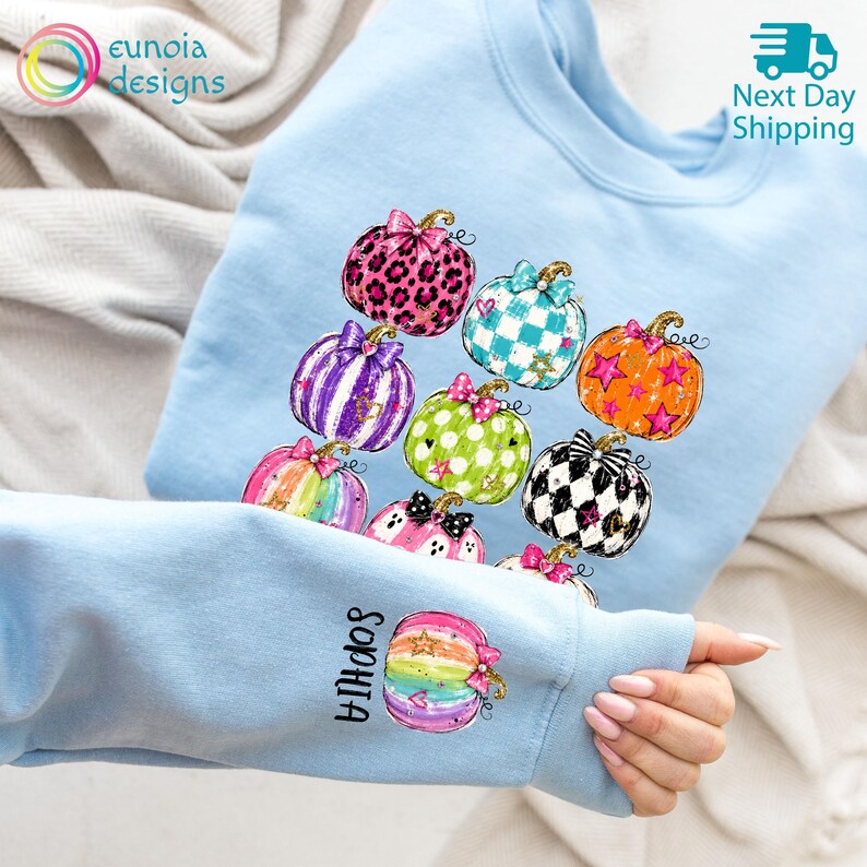 Puede incluir: Una sudadera azul claro con un estampado gr&aacute;fico de varias calabazas con diversos patrones, incluyendo estampado de leopardo, rayas, lunares y cuadros. La manga muestra una calabaza de colores arco&iacute;ris con el nombre Sophia impreso.