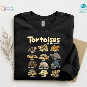 May include: Black sweatshirt featuring the text "Tortoises of the World" above illustrations of various tortoise species. The sweatshirt includes images of the Texas, Indian Star, Choco, Pancake, Spider, Galapagos, Red Footed, African Spurred, Aldabra, Radiated, Egyptian, and Leopard tortoises.