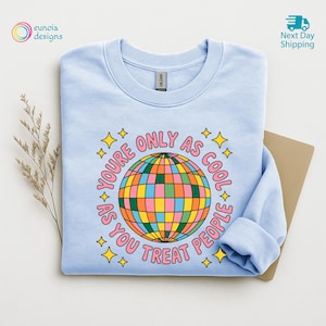 Op de afbeelding: Een lichtblauw sweatshirt met een kleurrijke discobal-afbeelding en de roze tekst "YOU'RE ONLY AS COOL AS YOU TREAT PEOPLE". Het ontwerp bevat gele sterren. Het sweatshirt is op een neutrale ondergrond gevouwen.