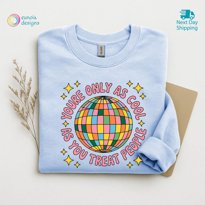 Op de afbeelding: Een lichtblauw sweatshirt met een kleurrijke discobal-afbeelding en de roze tekst "YOU'RE ONLY AS COOL AS YOU TREAT PEOPLE". Het ontwerp bevat gele sterren. Het sweatshirt is op een neutrale ondergrond gevouwen.