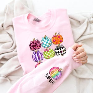 Puede incluir: Una sudadera de manga larga en color rosa claro con un estampado gr&aacute;fico de varias calabazas con diversos patrones, incluyendo estampado de leopardo, cuadros, estrellas y lunares. La manga muestra una calabaza arco&iacute;ris y el nombre Sophia en negro.