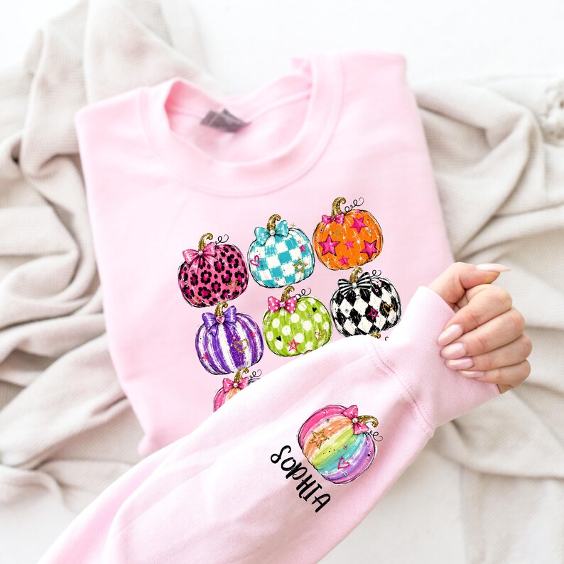 Puede incluir: Una sudadera de manga larga en color rosa claro con un estampado gr&aacute;fico de varias calabazas con diversos patrones, incluyendo estampado de leopardo, cuadros, estrellas y lunares. La manga muestra una calabaza arco&iacute;ris y el nombre Sophia en negro.