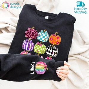 Puede incluir: Una sudadera negra de manga larga con un gr&aacute;fico de varias calabazas con diversos patrones, incluyendo estampado de leopardo, rayas, lunares y cuadros. La manga muestra una sola calabaza con el nombre Sophia impreso verticalmente.