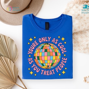 Op de afbeelding: Een koningsblauw t-shirt met de tekst "YOU'RE ONLY AS COOL AS YOU TREAT PEOPLE" rond een kleurrijk discobalontwerp. Het shirt is netjes opgevouwen op een witte ondergrond, met een hoed en decoratieve elementen op de achtergrond.
