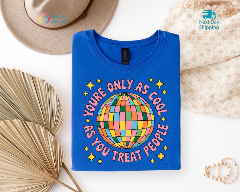 Op de afbeelding: Een koningsblauw t-shirt met de tekst "YOU'RE ONLY AS COOL AS YOU TREAT PEOPLE" rond een kleurrijk discobalontwerp. Het shirt is netjes opgevouwen op een witte ondergrond, met een hoed en decoratieve elementen op de achtergrond.