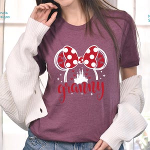 K&ouml;nnte beinhalten: Burgunderfarbenes T-Shirt mit einer wei&szlig;en Grafik eines roten und wei&szlig;en Bogens mit Mickey-Maus-Ohren und einem wei&szlig;en Schloss. Der Text "granny" befindet sich unterhalb der Grafik.