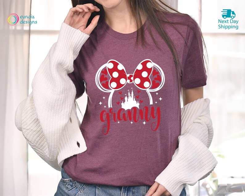 K&ouml;nnte beinhalten: Burgunderfarbenes T-Shirt mit einer wei&szlig;en Grafik eines roten und wei&szlig;en Bogens mit Mickey-Maus-Ohren und einem wei&szlig;en Schloss. Der Text "granny" befindet sich unterhalb der Grafik.