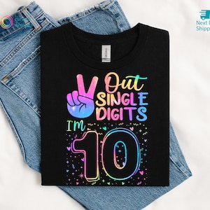 May include: Black t-shirt with the text "Out Single Digits I'm 10" in a rainbow gradient design. The design includes a peace sign hand gesture and the number 10. The shirt is displayed on a pair of blue jeans.