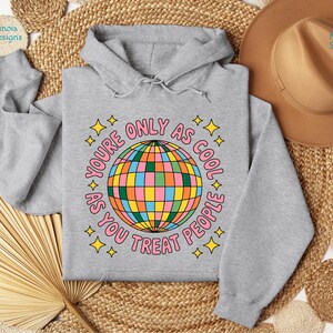 Op de afbeelding: Een lichtgrijze hoodie met een kleurrijk discobal-ontwerp. De tekst rond de discobal luidt "YOU'RE ONLY AS COOL AS YOU TREAT PEOPLE" in roze met gele sterren. De hoodie heeft een capuchon met trekkoord.