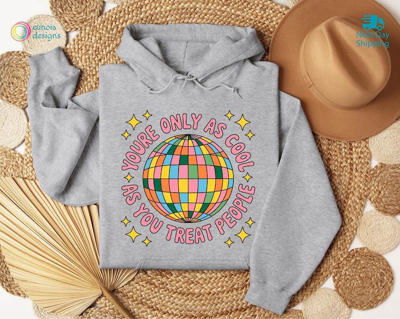 Op de afbeelding: Een lichtgrijze hoodie met een kleurrijk discobal-ontwerp. De tekst rond de discobal luidt "YOU'RE ONLY AS COOL AS YOU TREAT PEOPLE" in roze met gele sterren. De hoodie heeft een capuchon met trekkoord.