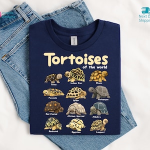 May include: A navy blue t-shirt featuring the text "Tortoises of the World" in gold lettering. Below the text are illustrations of various tortoise species, including Texas, Indian Star, and Galapagos. The shirt is displayed on a pair of blue jeans.