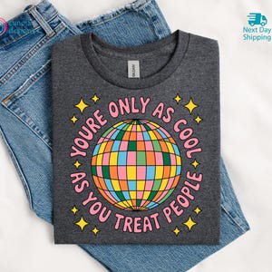 Op de afbeelding: Donkergrijs t-shirt met een kleurrijke discobal afbeelding en de tekst "You're only as cool as you treat people." Het t-shirt ligt op een blauwe spijkerbroek.