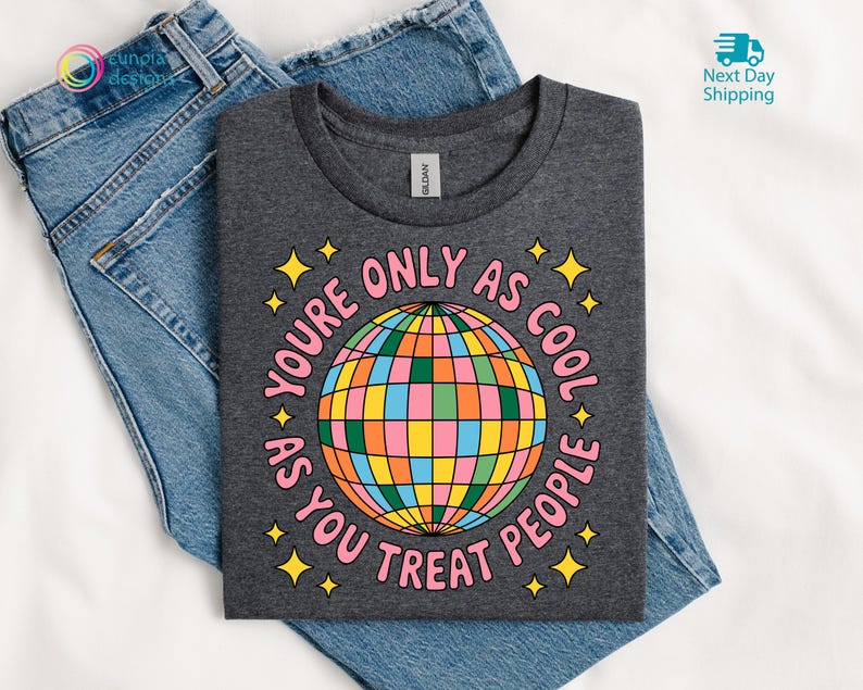 Op de afbeelding: Donkergrijs t-shirt met een kleurrijke discobal afbeelding en de tekst "You're only as cool as you treat people." Het t-shirt ligt op een blauwe spijkerbroek.
