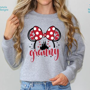 K&ouml;nnte beinhalten: Grauer Pullover mit einem roten und wei&szlig;en Polka-Dot-Minnie-Maus-Ohren-Design und dem Wort "granny" in roten Buchstaben. Die Ohren haben eine rote Schleife und eine schwarze Silhouette von Schloss Aschenputtel.