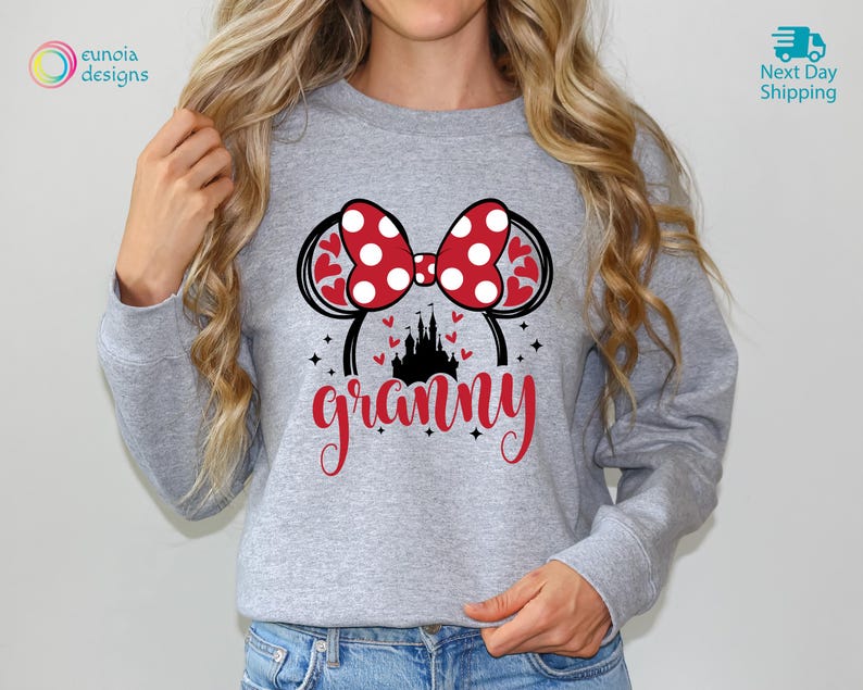 K&ouml;nnte beinhalten: Grauer Pullover mit einem roten und wei&szlig;en Polka-Dot-Minnie-Maus-Ohren-Design und dem Wort "granny" in roten Buchstaben. Die Ohren haben eine rote Schleife und eine schwarze Silhouette von Schloss Aschenputtel.