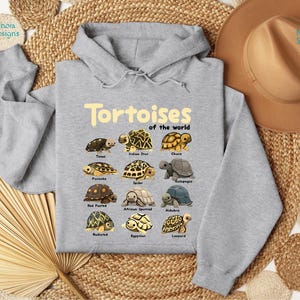 May include: A light gray hooded sweatshirt featuring the text "Tortoises of the World" in a yellow font. Below the text are illustrations of various tortoise species. The sweatshirt is displayed on a woven surface with a brown hat.