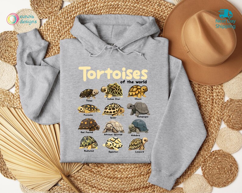 May include: A light gray hooded sweatshirt featuring the text "Tortoises of the World" in a yellow font. Below the text are illustrations of various tortoise species. The sweatshirt is displayed on a woven surface with a brown hat.