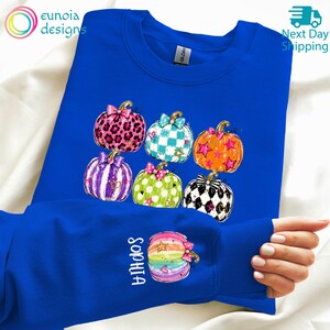 Puede incluir: Una sudadera azul real de cuello redondo con un estampado gr&aacute;fico de seis calabazas con varios patrones, incluyendo estampado de leopardo, cuadros y estrellas. La manga muestra una calabaza de colores arco&iacute;ris con el nombre Sophia en blanco.