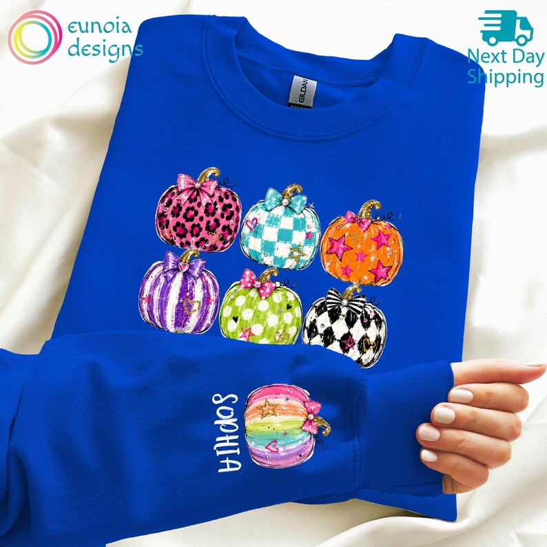 Puede incluir: Una sudadera azul real de cuello redondo con un estampado gr&aacute;fico de seis calabazas con varios patrones, incluyendo estampado de leopardo, cuadros y estrellas. La manga muestra una calabaza de colores arco&iacute;ris con el nombre Sophia en blanco.
