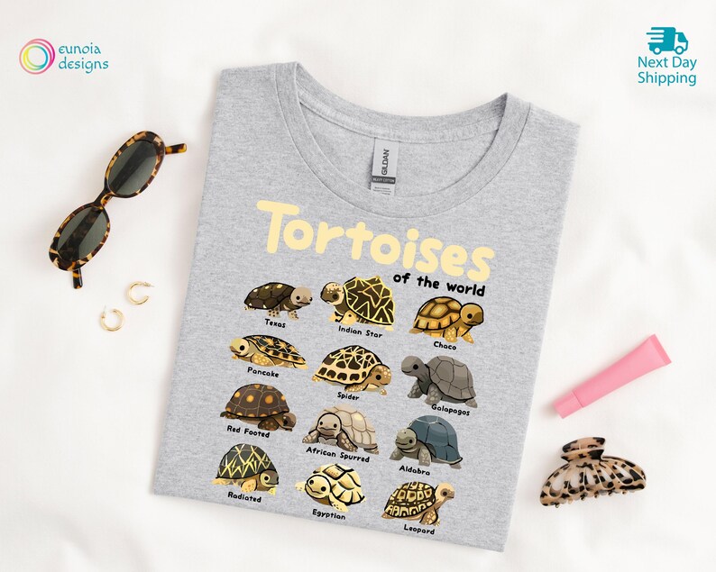 May include: A light gray t-shirt featuring the text "Tortoises of the World" in yellow, with illustrations of various tortoise species. The shirt is surrounded by accessories, including sunglasses, earrings, and a hair clip.
