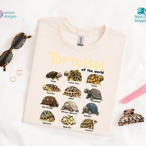 May include: Cream-colored t-shirt featuring the text "Tortoises of the World" in yellow, with illustrations of various tortoise species. The shirt includes a crew neck and is made of a soft material.