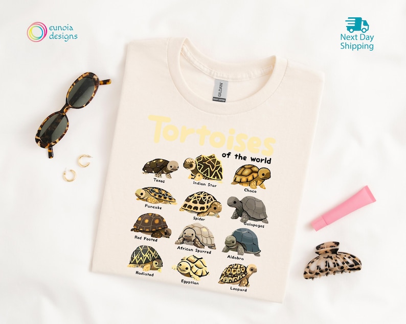 May include: Cream-colored t-shirt featuring the text "Tortoises of the World" in yellow, with illustrations of various tortoise species. The shirt includes a crew neck and is made of a soft material.