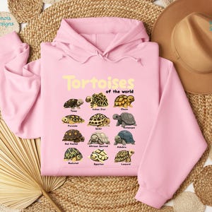 May include: A light pink hoodie featuring the text "Tortoises of the world" in yellow, along with illustrations of various tortoise species. The hoodie has a drawstring hood and long sleeves. The design includes species such as Texas, Indian Star, and Galapagos.
