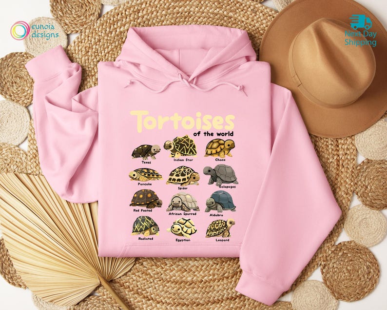 May include: A light pink hoodie featuring the text "Tortoises of the world" in yellow, along with illustrations of various tortoise species. The hoodie has a drawstring hood and long sleeves. The design includes species such as Texas, Indian Star, and Galapagos.