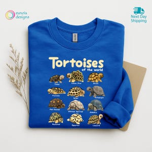 May include: A royal blue sweatshirt featuring the text "Tortoises of the World" above illustrations of various tortoise species. The sweatshirt has a crew neck and long sleeves. The brand name "Gildan" is visible.