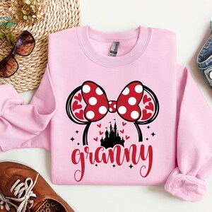 K&ouml;nnte beinhalten: Rosafarbener Pullover mit einem roten und wei&szlig;en Minnie Mouse-Ohren-Design, einer schwarzen Schlossgrafik und dem Wort "granny" in roten Buchstaben.