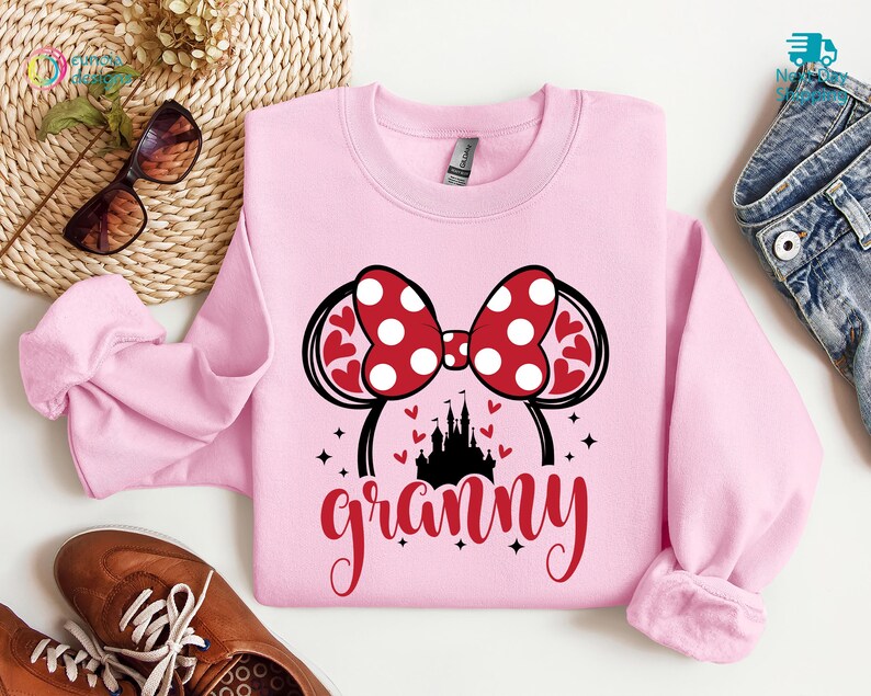 K&ouml;nnte beinhalten: Rosafarbener Pullover mit einem roten und wei&szlig;en Minnie Mouse-Ohren-Design, einer schwarzen Schlossgrafik und dem Wort "granny" in roten Buchstaben.