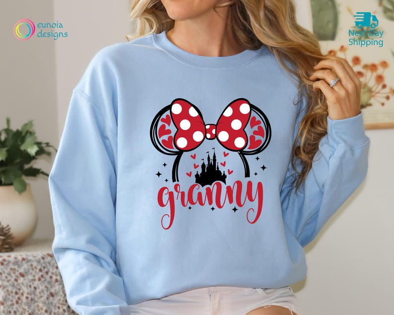 K&ouml;nnte beinhalten: Hellblauer Pullover mit einem roten und wei&szlig;en Minnie Mouse Ohren-Design und dem Wort "grammy" in roten und schwarzen Buchstaben.