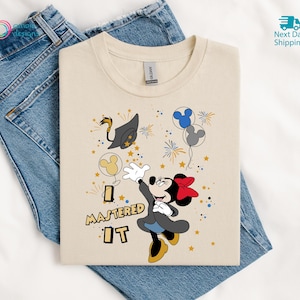 May include: A cream-colored t-shirt featuring Minnie Mouse celebrating graduation. The design includes a graduation cap, balloons, fireworks, and the text "I MASTERED IT" in a playful font. The shirt is laid on top of blue jeans.