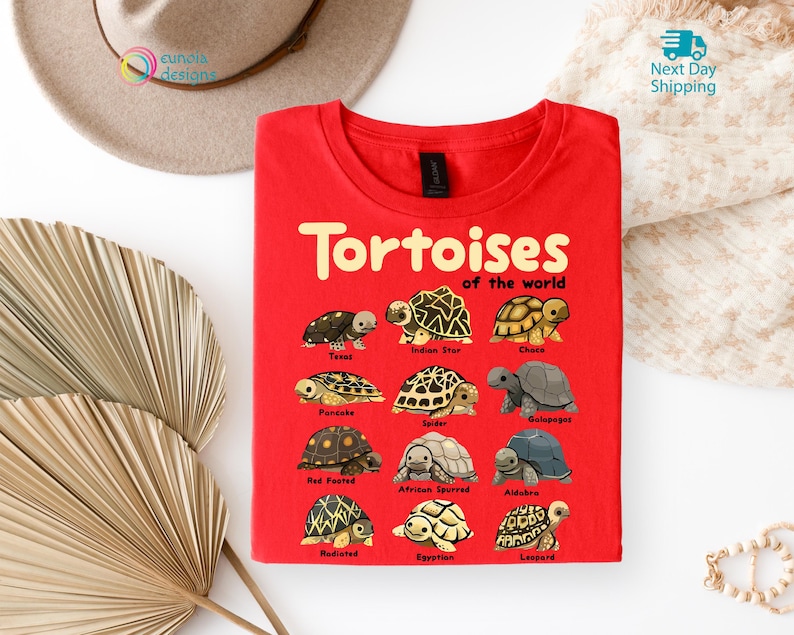 May include: A red t-shirt featuring the text "Tortoises of the World" in yellow, with illustrations of various tortoise species. The shirt includes names like Texas, Indian Star, and Chaco. The design is centered on the front of the shirt.
