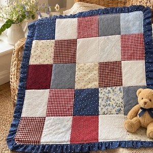 May include: A patchwork quilt featuring a grid of squares in red, white, and blue patterns, including gingham, floral, and polka dot prints. The quilt is finished with a dark blue ruffled border with white dots and includes a small teddy bear.