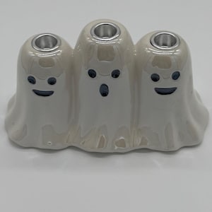 May include: A glossy off-white ceramic candle holder shaped like three conjoined ghosts. Each ghost figure features dark blue painted eyes and a mouth, with a circular silver-toned metal insert at the top of each head for holding candles.