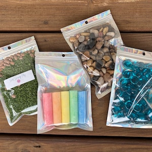 May include: Four clear, iridescent zip-lock bags filled with various sensory items. One bag contains green dyed rice and beans with a label that reads "Soft Sensory Play". Other bags hold smooth stones, colorful chalk sticks, and blue glass gems.