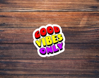 May include: A white sticker with the words "GOOD VIBES ONLY" in colorful, outlined letters. The letters are red, yellow, and purple. The sticker is on a brown wooden background.