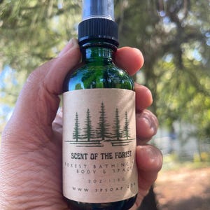 May include: A green glass spray bottle with a black nozzle and a tan paper label. The label features a black graphic of pine trees and the text: SCENT OF THE FOREST, FOREST BATHING, BODY & SPACE, 2 OZ / 118 G, WWW.3PSOAP.COM. The bottle contains liquid.