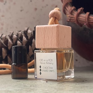 May include: A small glass bottle with a black cap next to a larger, clear glass bottle with a wooden top. The larger bottle has a label that reads "3PS in a POD Native Alchemy CHOCTAW KINDRED SPIRITS". A beige cord and wooden bead are attached to the top.
