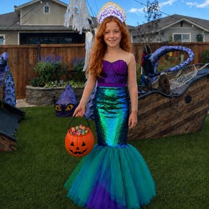 May include: A mermaid costume featuring a purple sequined bodice with a sweetheart neckline, long sheer sleeves, and a green and blue sequined skirt with a layered tulle hem. The outfit includes a white and gold crown and an orange pumpkin bucket.