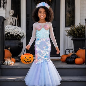 May include: A mermaid costume featuring a light blue bodice with iridescent shell accents, long sheer sleeves, and a skirt covered in shimmering, scale-like sequins that transitions into a layered, light purple tulle hem, paired with a matching crown.