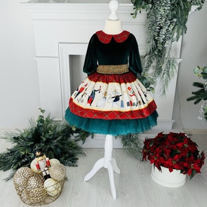 Puede incluir: Un vestido de terciopelo verde con cuello blanco y falda roja y blanca con una escena del Cascanueces. La falda tiene una capa de tul y un cinturón dorado.