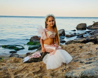 May include: A young girl wearing a rose gold mermaid costume with a white tulle skirt and a white headband. She is sitting on rocks by the ocean.